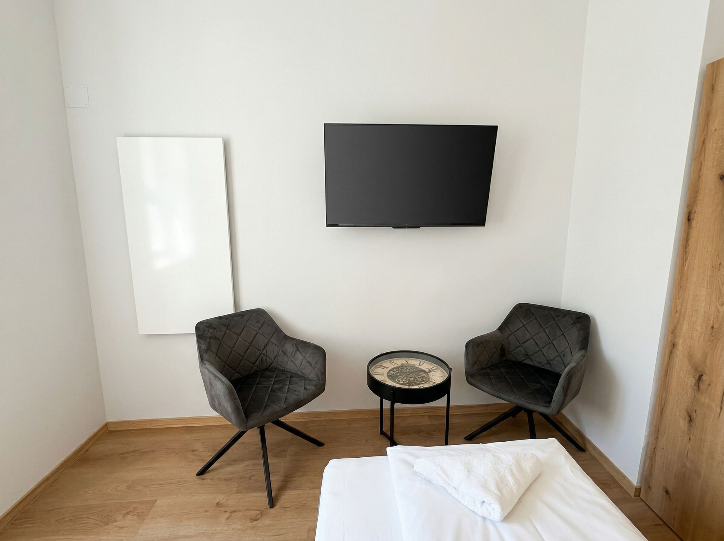 Ett hotellrum med en säng, två fåtöljer, ett runt sidobord med en klocka och en platt-TV som är monterad på väggen.
