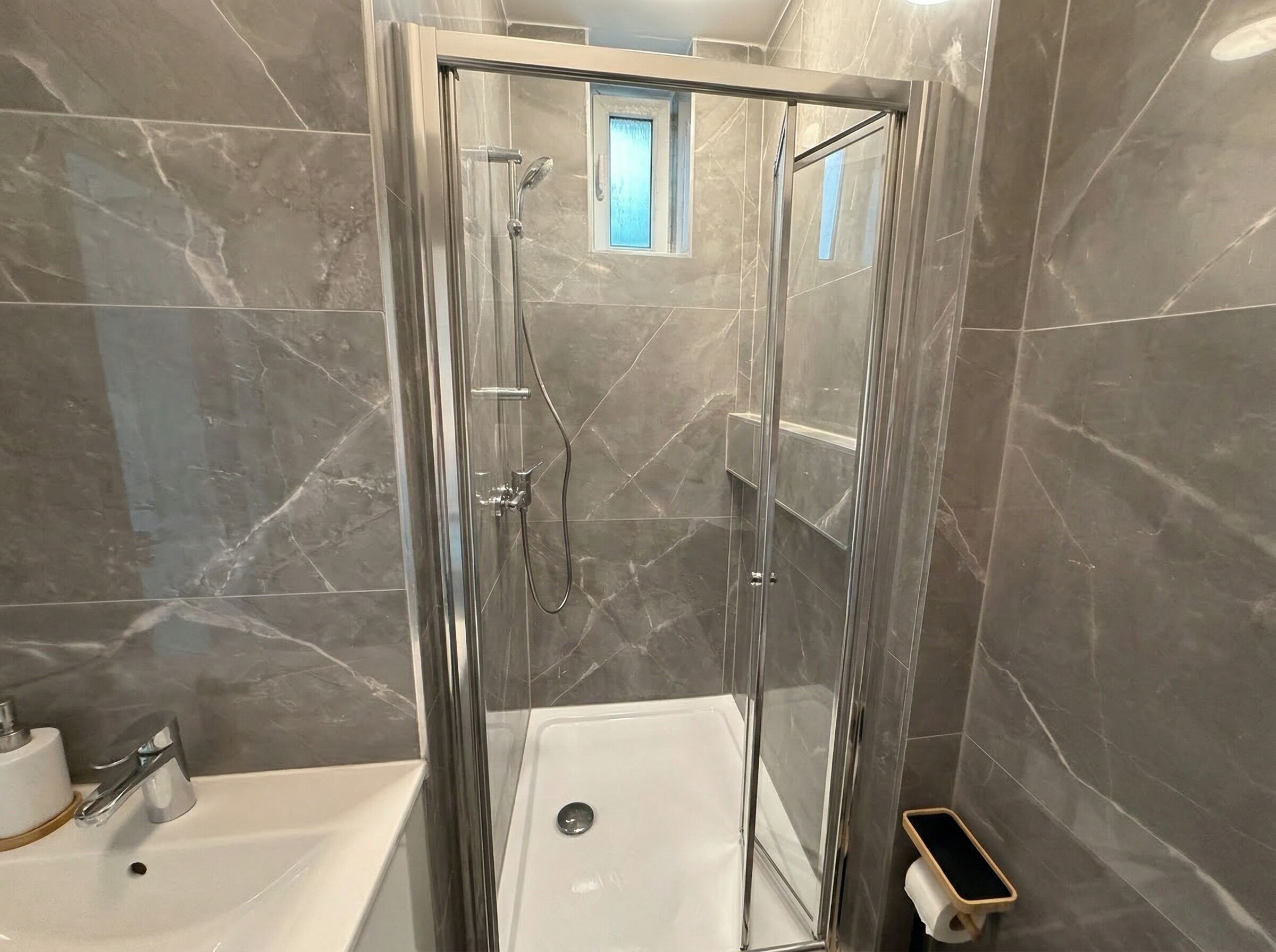Een moderne badkamer met een glazen douchecabine, marmeren tegels, een witte badkuip en een klein raam.