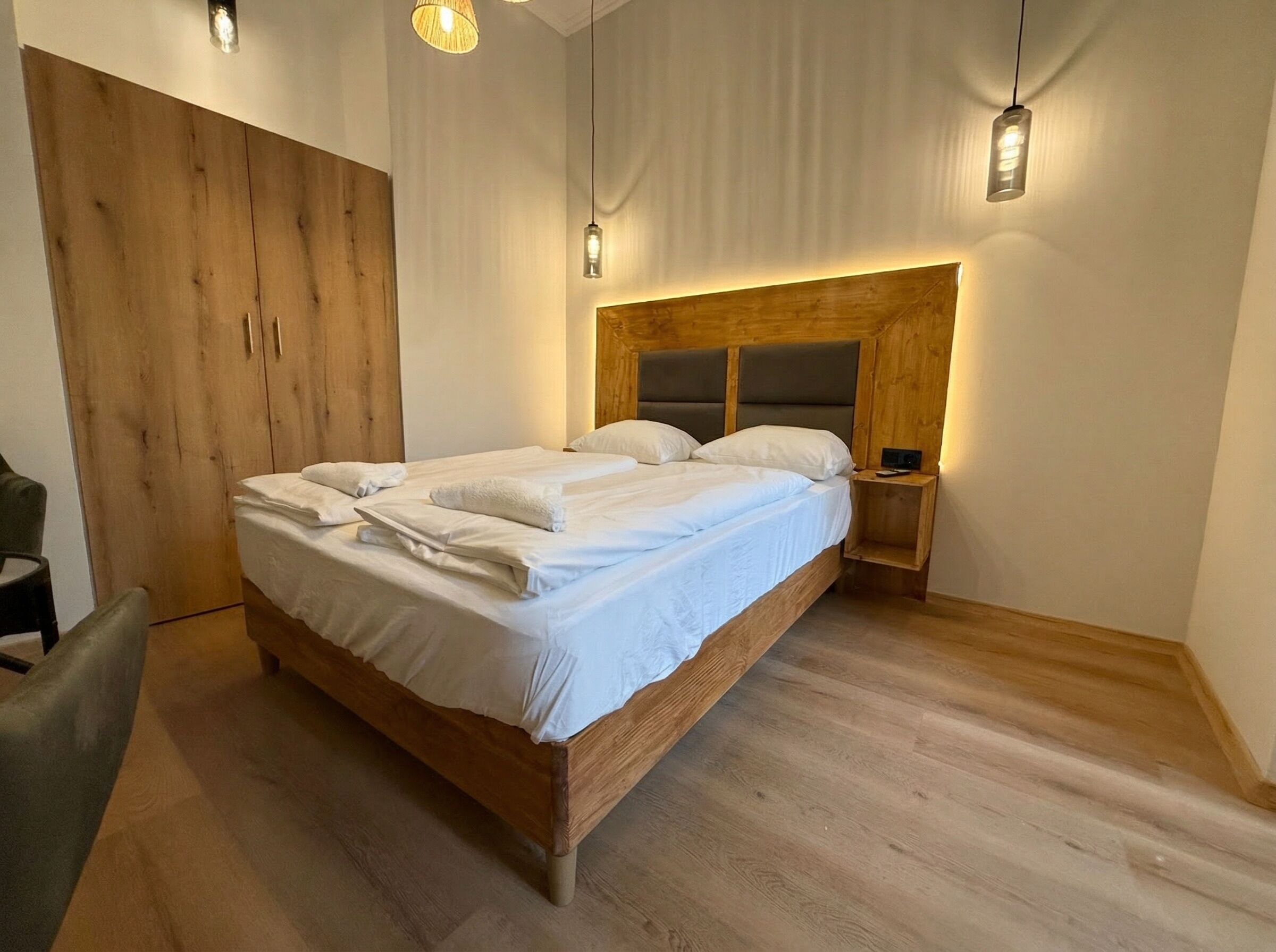 A neatly made bed with white linens and pillows, a wooden wardrobe, a bedside table with a lamp, and a wooden floor.