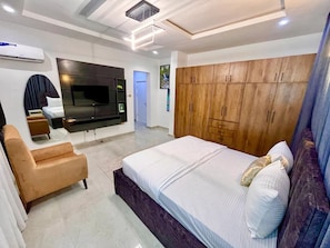 A modern bedroom with a large bed, a TV mounted on the wall, a wooden wardrobe, and a seating area with a sofa and armchair. - Red Box Apartment (Port Harcourt)
