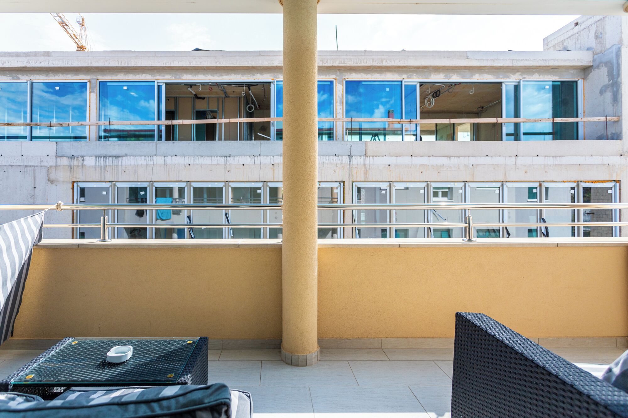 Un balcón de hotel con vistas a un edificio de múltiples ventanas y fachada de hormigón. Hay dos sillas de mimbre y una pequeña mesa con una taza encima.