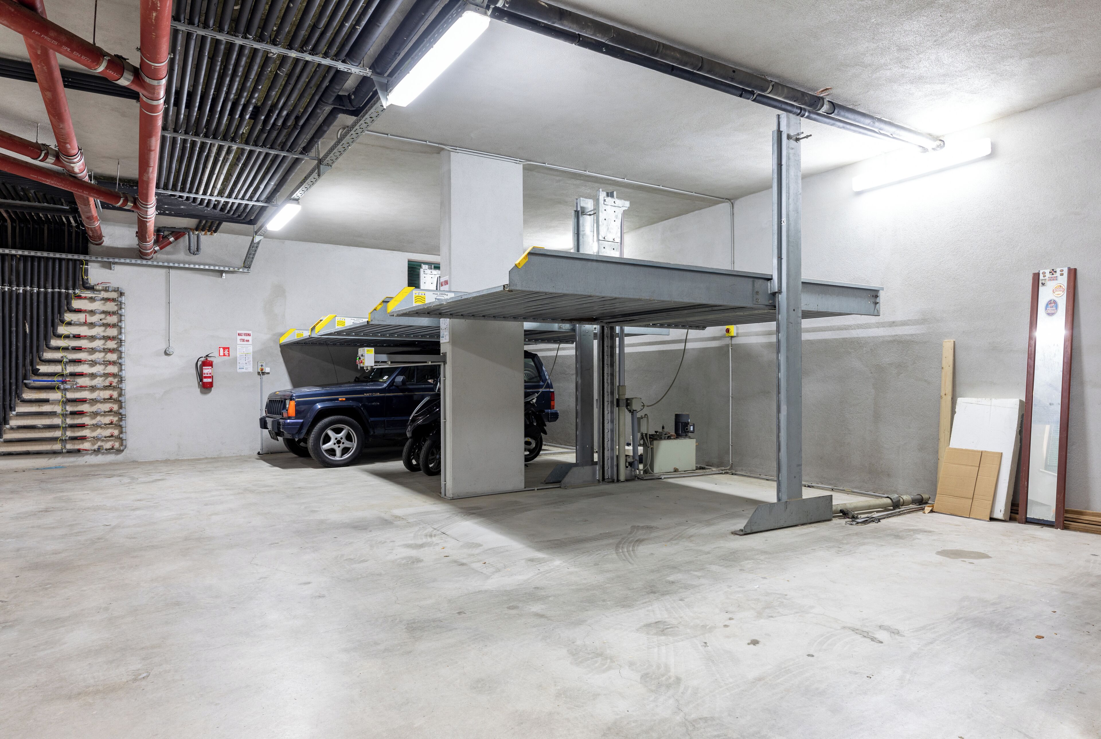 Un parcheggio con un'auto blu sotto una tettoia, pavimenti in cemento e tubature a vista sul soffitto.