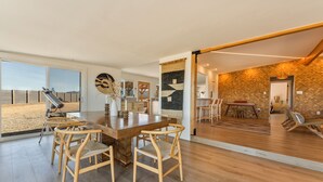 A spacious living area with a wooden dining table and chairs, a large window offering a view of the outdoors, and a cozy seating area with a hammock. - Stylish Desert Retreat - Hot Tub, Game Room, Fire Pit, Minutes to JTNP (Twentynine Palms)