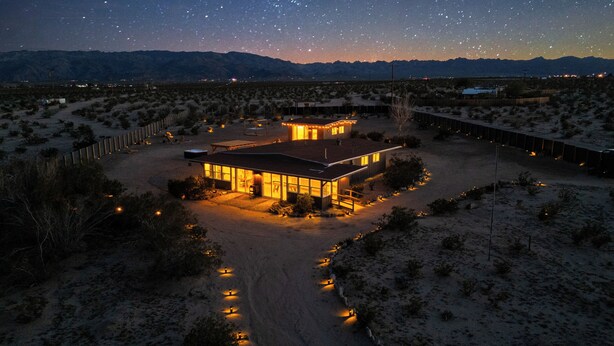 A modern house with a large patio, surrounded by desert vegetation, under a starry night sky. - Stylish Desert Retreat - Hot Tub, Game Room, Fire Pit, Minutes to JTNP (Twentynine Palms)