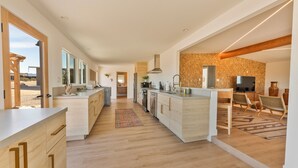 A modern kitchen with wooden cabinets, a central island, and a dining area with wicker chairs. - Stylish Desert Retreat - Hot Tub, Game Room, Fire Pit, Minutes to JTNP (Twentynine Palms)