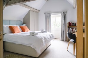 A bedroom with a bed, pillows, a bedside table, a window with curtains, a bookshelf, and a chair. - Stunning 6 Bedroom Riverside Barn Cotswold Mill (Cirencester)