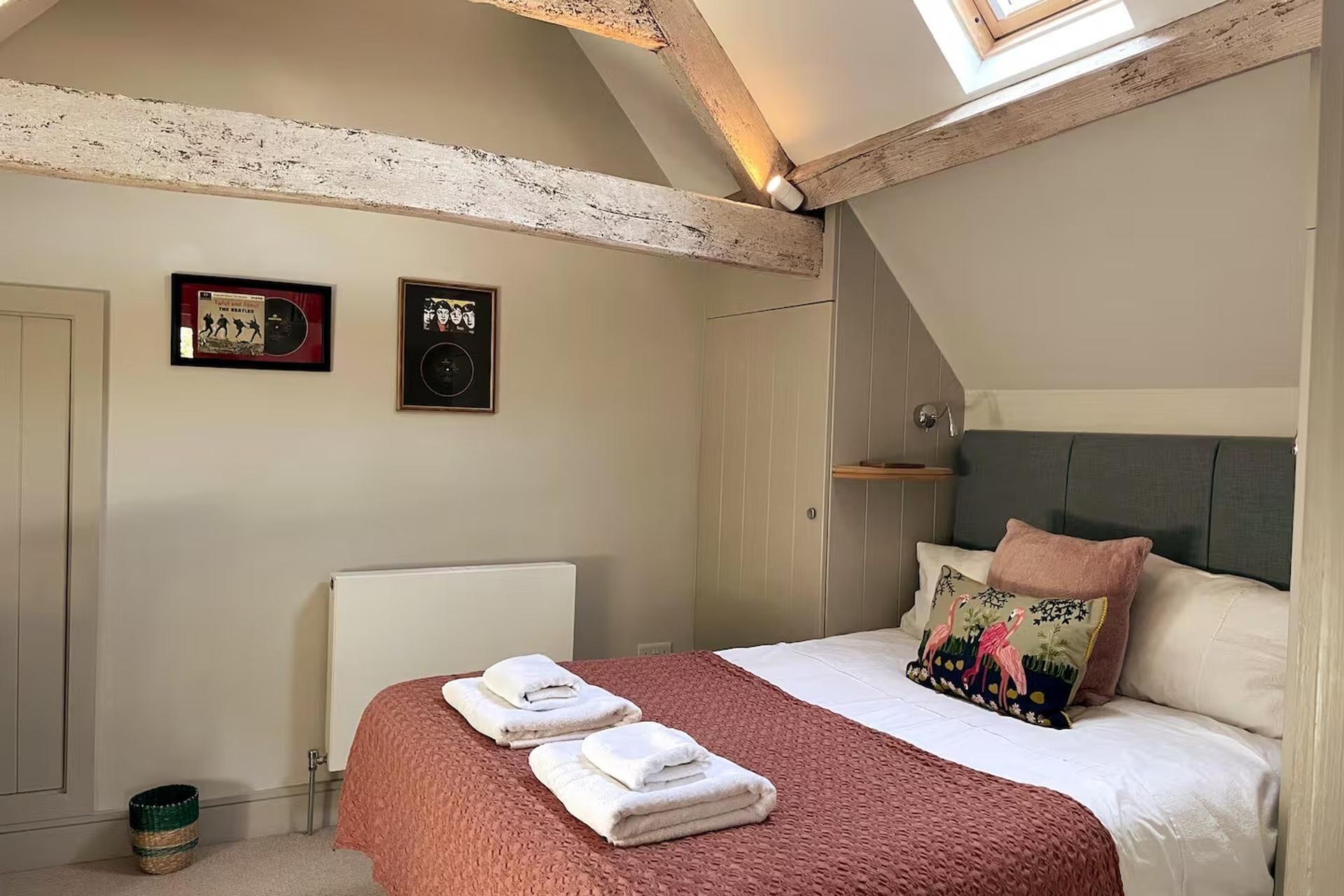 A bedroom with a sloped ceiling, a bed with a pink blanket, a radiator, and two framed posters on the wall.