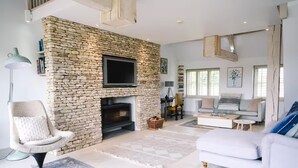 A modern living room with a stone fireplace, a flat-screen TV, a white sofa, and a reading chair. - Stunning 6 Bedroom Riverside Barn Cotswold Mill (Cirencester)