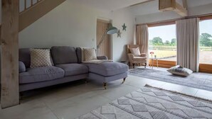 A living room with a grey sectional sofa, a small round table, and a chair. There is a staircase on the left, a window with curtains, and a view of a field outside. - Stunning 6 Bedroom Riverside Barn Cotswold Mill (Cirencester)