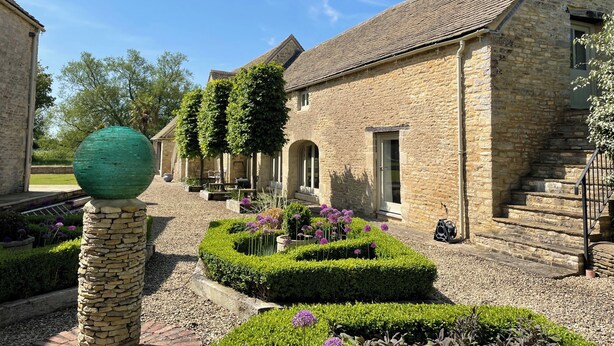 A stone building with a tiled roof, a stone pillar, and a well-maintained garden with hedges and purple flowers. - Stunning 6 Bedroom Riverside Barn Cotswold Mill (Cirencester)
