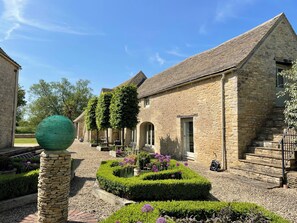 A stone building with a tiled roof, a stone pillar, and a well-maintained garden with hedges and purple flowers. - Stunning 6 Bedroom Riverside Barn Cotswold Mill (Cirencester)