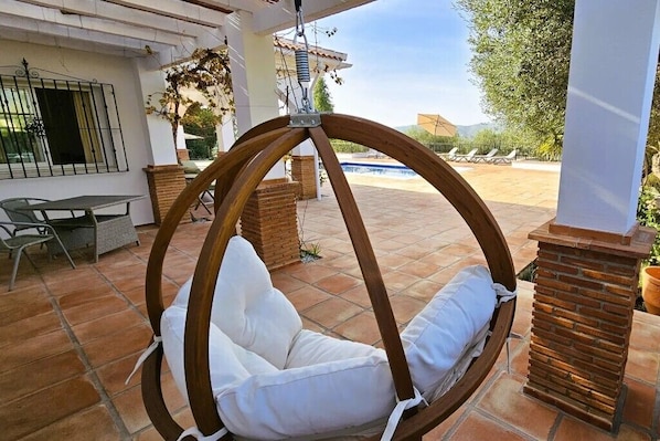 A wooden swing chair with white cushions in a covered outdoor area with tiled flooring, brick pillars, and a view of a swimming pool and greenery.