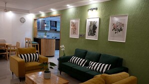 Een woonkamer met een groene wand, een groene bank, een geel fauteuil en een houten salontafel. Op de wand hangt een klok en boven de bank is er kunst in lijsten.