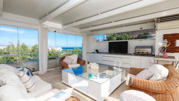 A modern living room with a glass coffee table, white sofas, wicker chairs, a TV, and a kitchen area with a grill.