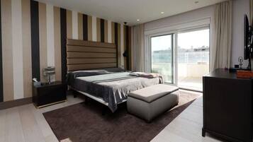 A modern bedroom with a large bed, a nightstand, a dresser, and a view of the city through a sliding glass door.