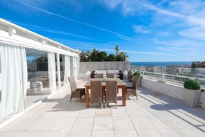 Een terras op het dak met een eettafel en stoelen, een glazen deur naar een zithoek, en uitzicht op zee.