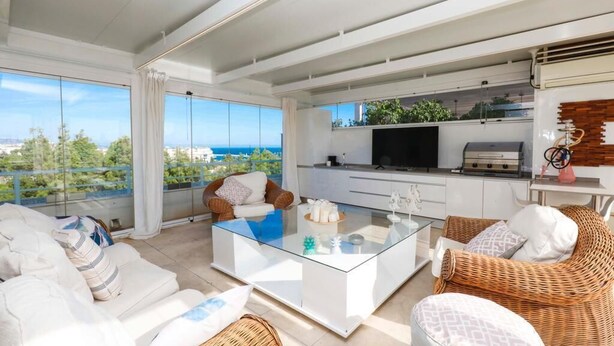 Un salon moderne avec une table basse en verre, des canapés blancs, des fauteuils en osier, un téléviseur et un coin cuisine équipé d’un barbecue.