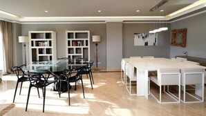 Un espace repas moderne avec une table à plateau en verre, des chaises noires et des bancs blancs. Deux étagères blanches sont adossées au mur, et chacune est surmontée d’une lampe.