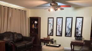 Ein Wohnzimmer mit dunklem Holzboden, einem Ledersofa, einem runden Hocker, einem Couchtisch und einem Deckenventilator. An der Wand befinden sich gerahmte Kunstwerke und ein kleiner Beistelltisch mit einer Vase.