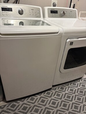 Vista frontal de una lavadora y secadora Samsung blancas de carga frontal, con panel de control, puerta y alfombra estampada en el suelo.