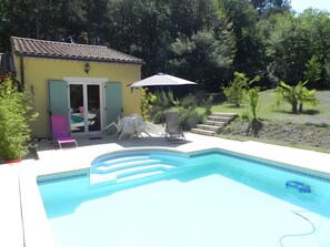 Une piscine avec un espace patio, un petit bâtiment doté d’une porte et des marches menant à une zone herbeuse.