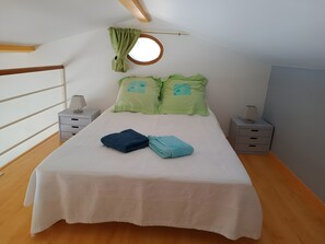 Un lit bien fait, recouvert d’une couvre-lit blanche, avec deux oreillers à motifs verts et deux serviettes pliées. On y trouve deux tables de chevet avec des lampes, une fenêtre offrant une vue circulaire et un plancher en bois.