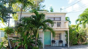 Una casa de dos pisos con balcón, rodeada de plantas tropicales y un camino de grava.