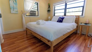 Un dormitorio con cama de madera, ropa de cama blanca, una mesita de noche con lámpara y una ventana con persianas.