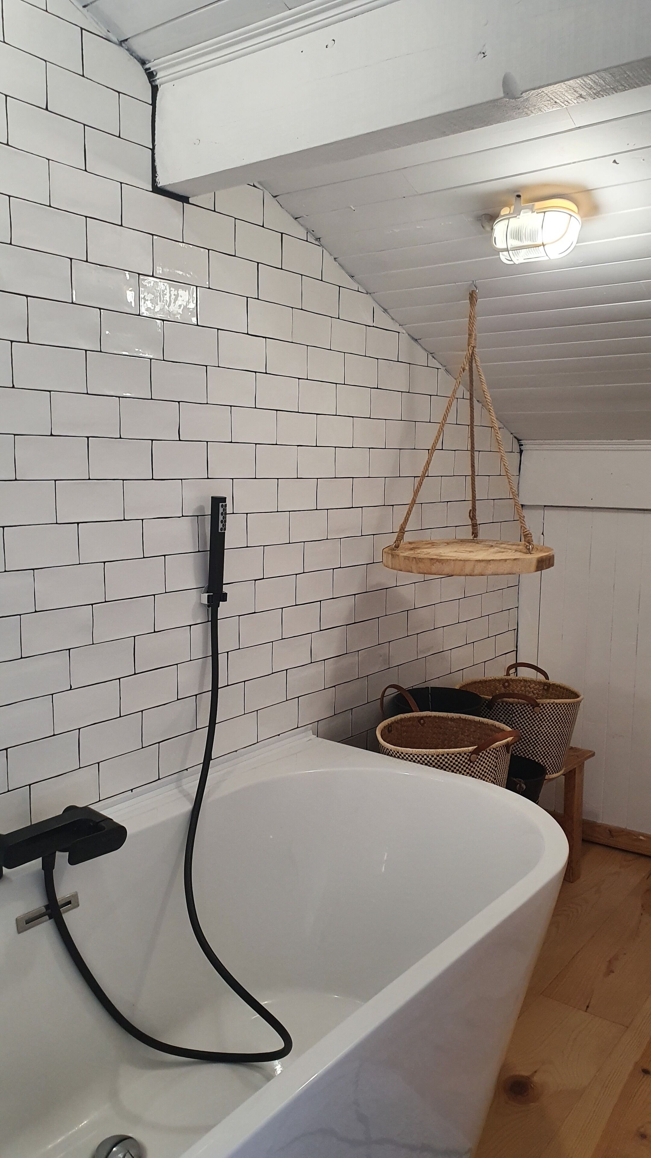 A bathroom with a white bathtub, a hanging wooden shelf, and a white tiled wall.