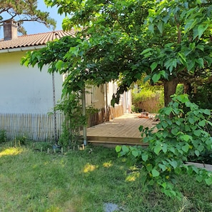A residential backyard with a wooden deck, a white house, and a tree providing shade. - La maison des muriers in a peaceful setting (Mimizan)