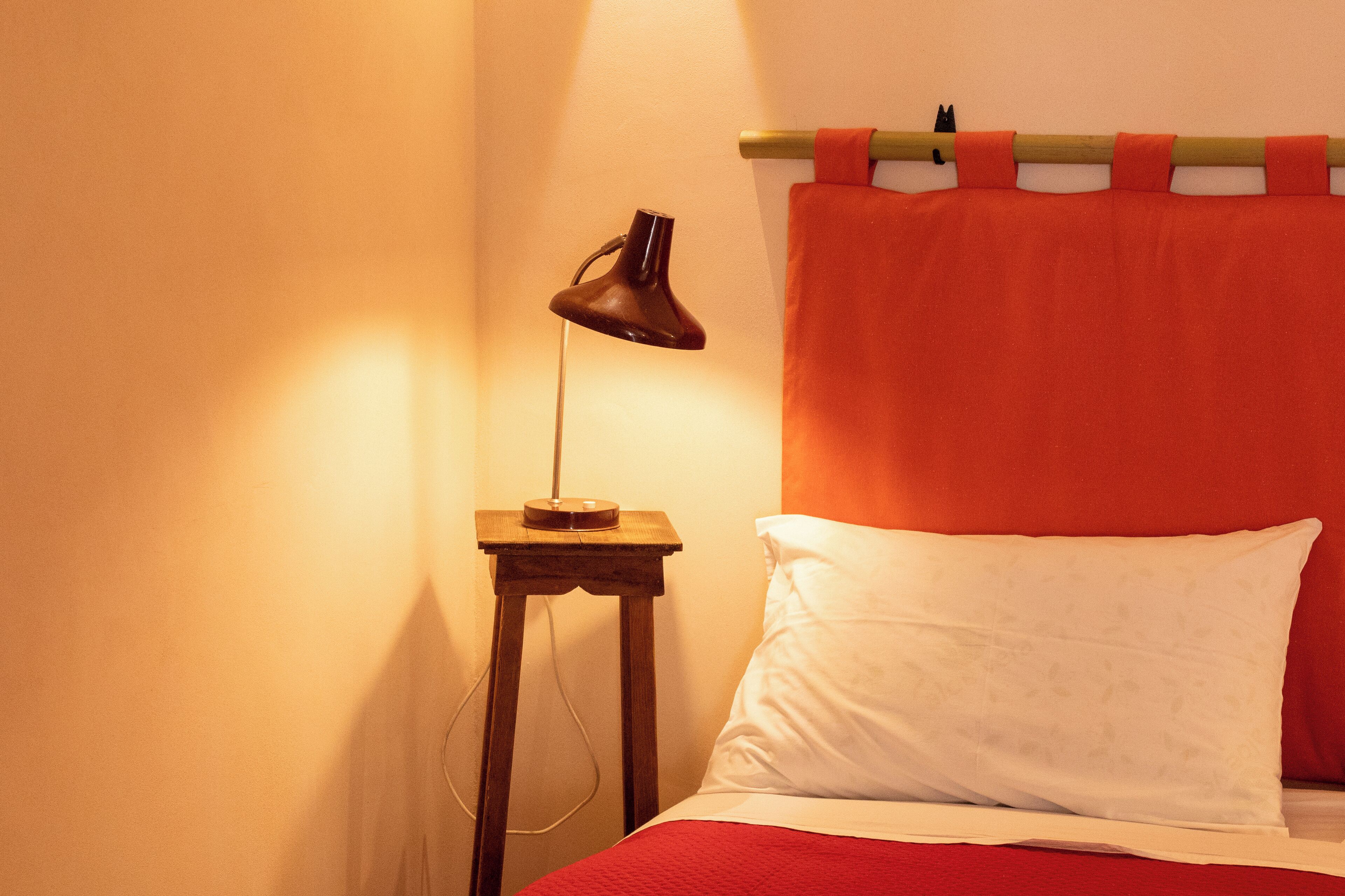 A bed with a red blanket, a white pillow, and a wooden bedside table with a lamp.