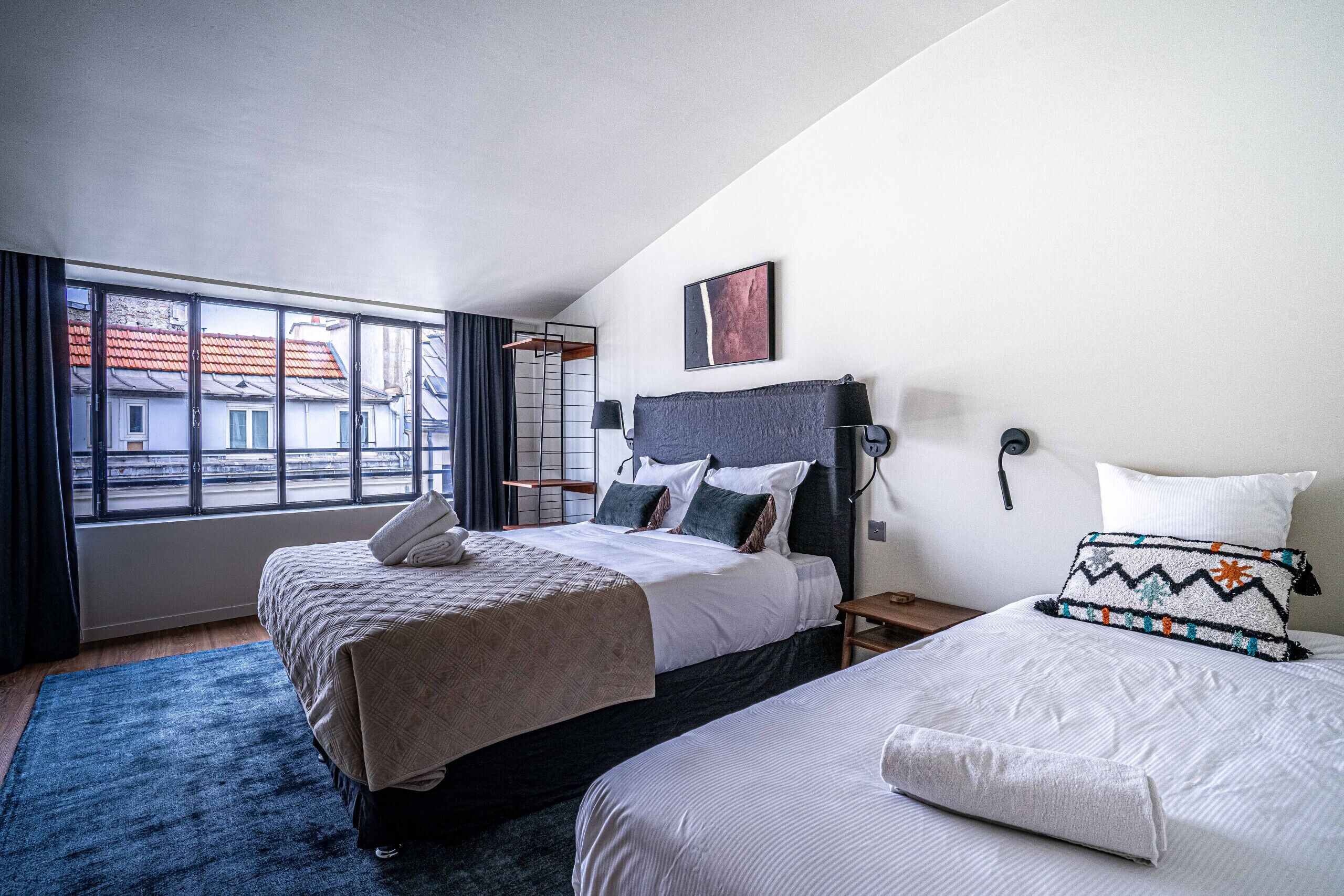 A hotel room with two beds, a large window with a view of buildings, a black headboard, and a wall-mounted lamp.
