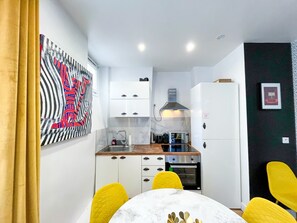 A compact kitchen with white cabinets, a microwave, and a stove. There is a yellow chair and a table with a white tablecloth. - 158 - Urban Louis Flat in Montorgueil (París)