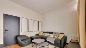 Ein modernes Wohnzimmer mit einem dunkelgrauen Sofa, einem runden Couchtisch und einem grauen Sessel. An der Wand hängen vier gerahmte Kunstwerke, und die Rückwand ist mit einem gemusterten Wandpapier gestaltet.