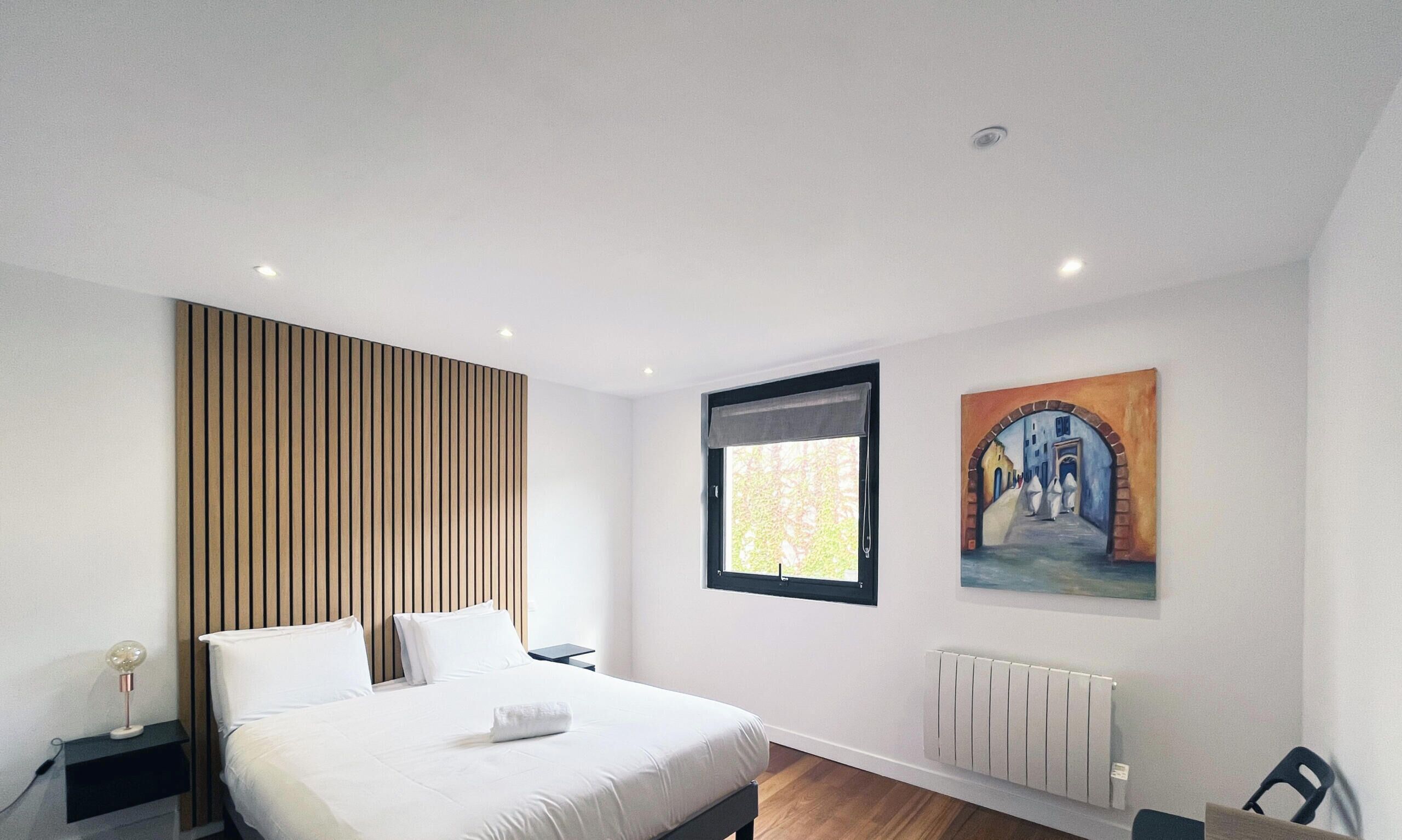 A modern bedroom with a bed, a nightstand, a window with a view, a radiator, and a painting on the wall.