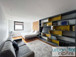 Un dormitorio moderno con una cama, un sofá gris, una estantería y una ventana.