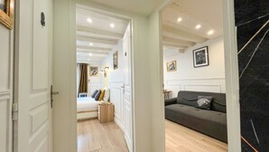 Un pasillo que conduce a un dormitorio con una cama, una mesita de noche y un aparador. En la habitación contigua hay un sofá de color oscuro.
