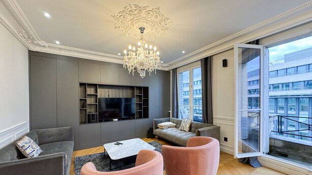 Ein modernes Wohnzimmer mit Flachbildfernseher, grauen Sofas, einer Kristallleuchtre und großen Fenstern mit Blick auf die Stadt.