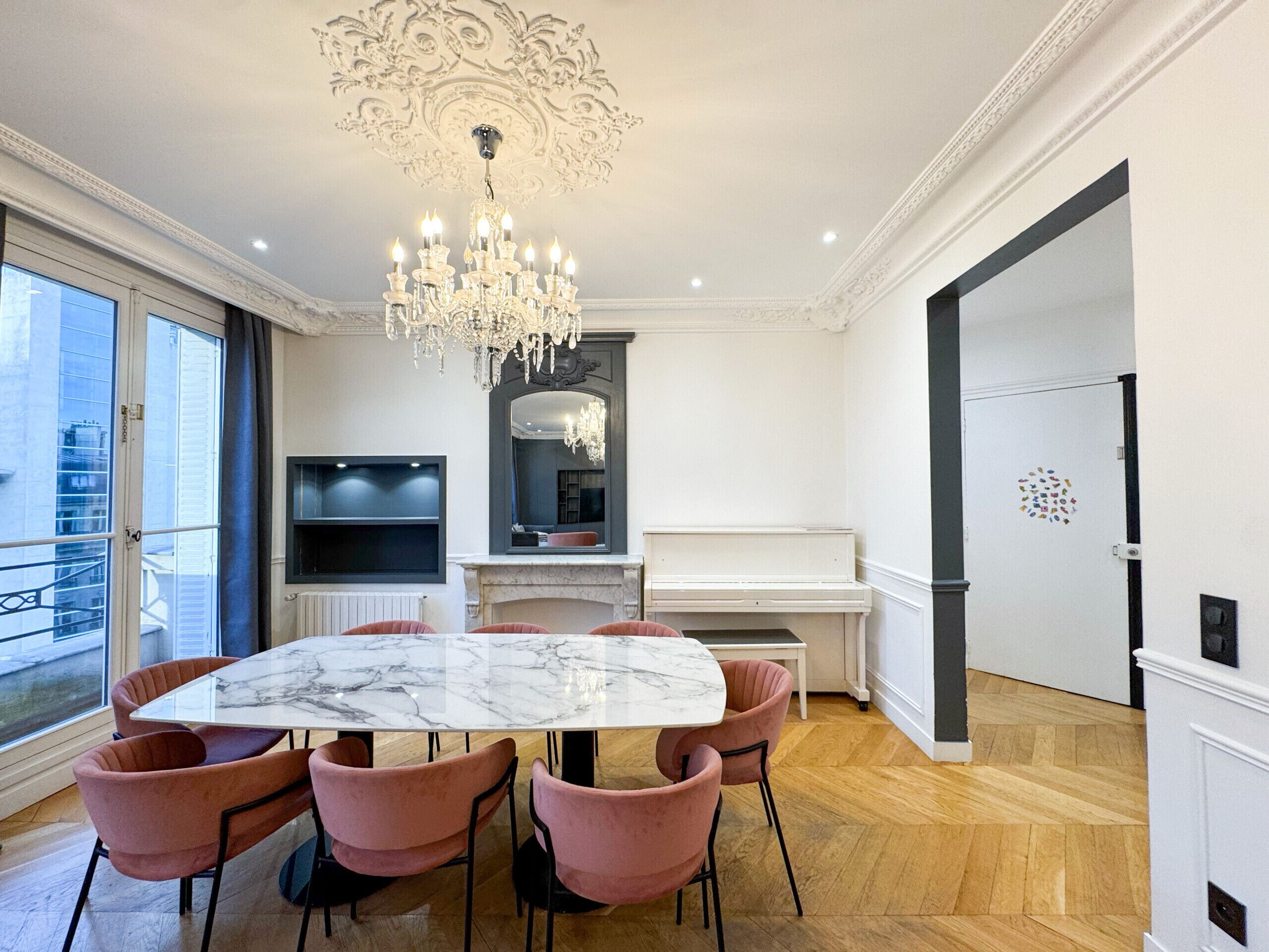 Een moderne eetkamer met een marmeren tafel, roze stoelen, een kroonluchter, een televisie, een open haard en een piano.