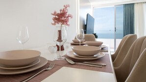 Ein moderner Essbereich mit einem weißen Tisch, gedeckt für vier Personen, mit beigen Tellern und Gläsern, einer Vase mit rosa Blumen und Blick auf das Meer durch große Fenster.