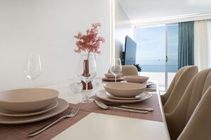 Ein moderner Essbereich mit einem weißen Tisch, gedeckt für vier Personen, mit beigen Tellern und Gläsern, einer Vase mit rosa Blumen und Blick auf das Meer durch große Fenster.