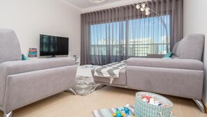 Een moderne woonkamer met twee grijze banken, een flatscreen televisie, een groot raam met gordijnen en een plafondlamp.
