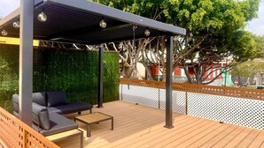 Ein überdachter Sitzbereich im Freien mit einer Couch und einem kleinen Tisch, umgeben von einer Holzterrasse und einer grünen Wand.