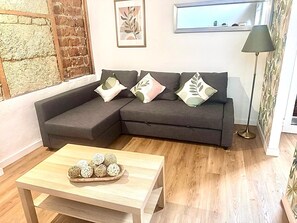 Un salon moderne avec un canapé d’angle, une table basse en bois agrémentée d’objets décoratifs, un lampadaire et un mur en briques orné d’un tableau encadré.