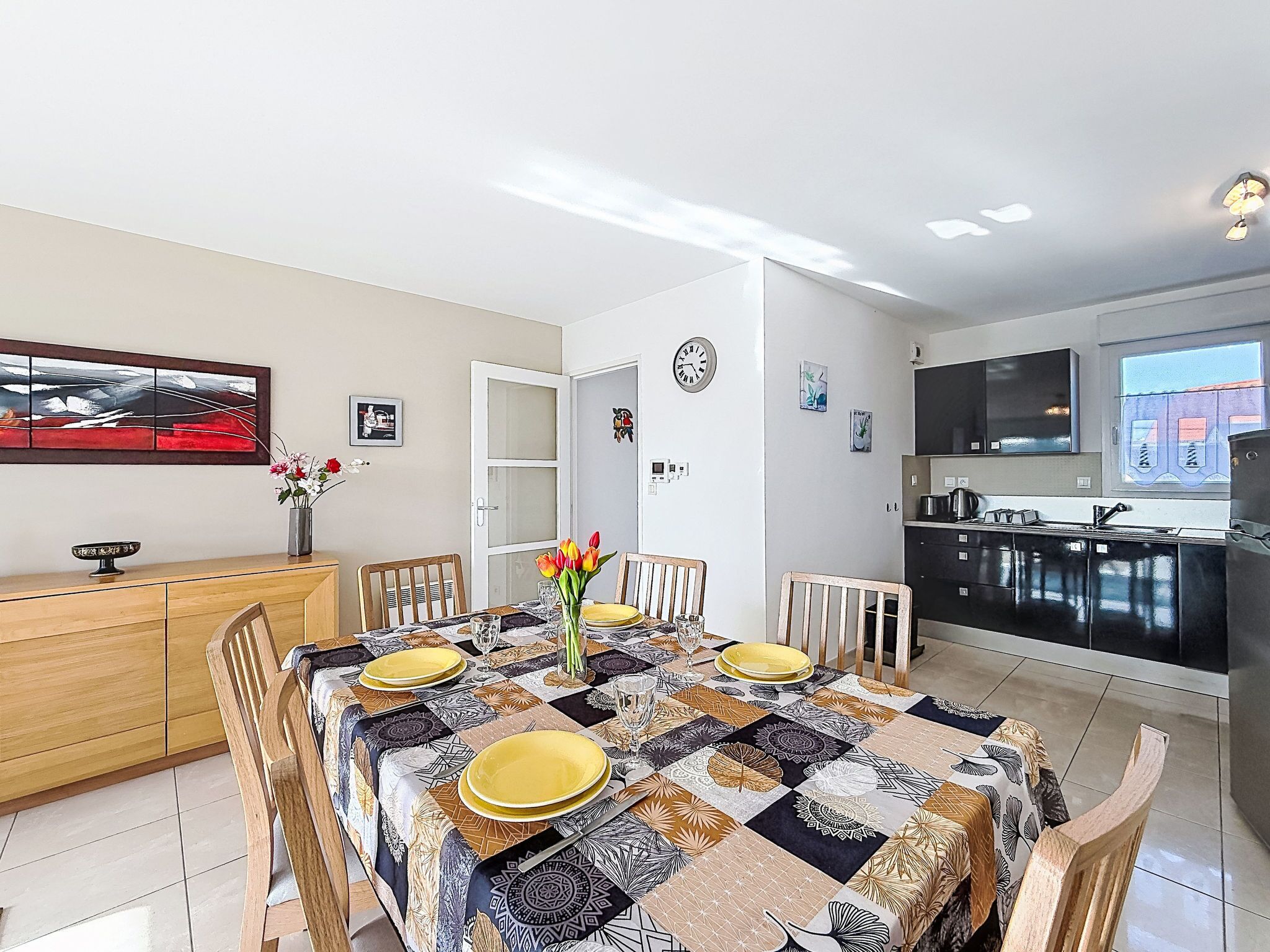 Un coin repas avec une table dressée pour quatre personnes, une cuisine en arrière-plan et une porte donnant sur une autre pièce.