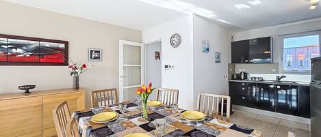 Un coin repas avec une table dressée pour quatre personnes, une cuisine en arrière-plan et une porte donnant sur une autre pièce.