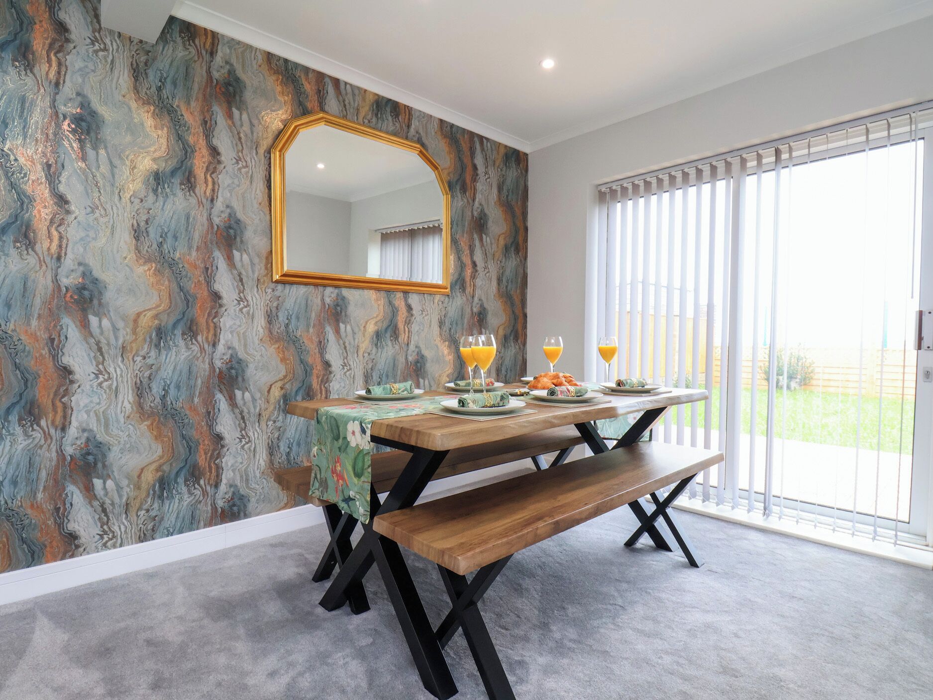 Un comedor con una pared con diseño de mármol, una mesa de madera con bancos y un amplio ventanal con persianas.