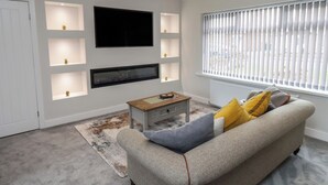 Ein Wohnzimmer mit einem an der Wand befestigten Flachbildfernseher, einem Holztisch, einem grauen Sofa mit gelb-blauen Kissen und einem Fenster mit vertikalen Jalousien.