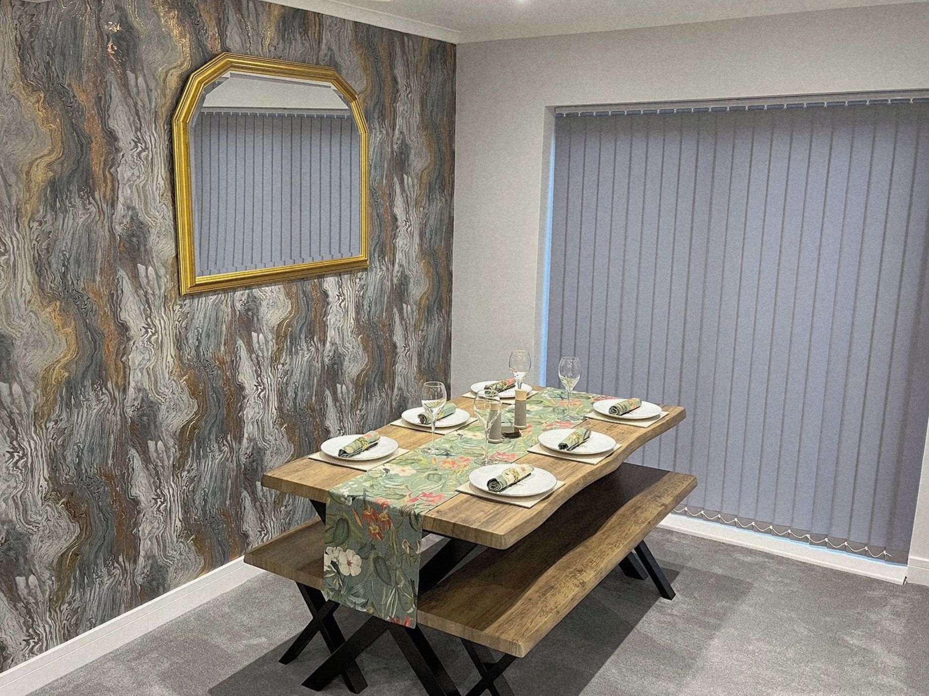 A dining area with a wooden table set for four, a mirror on the wall, and a window with blinds.