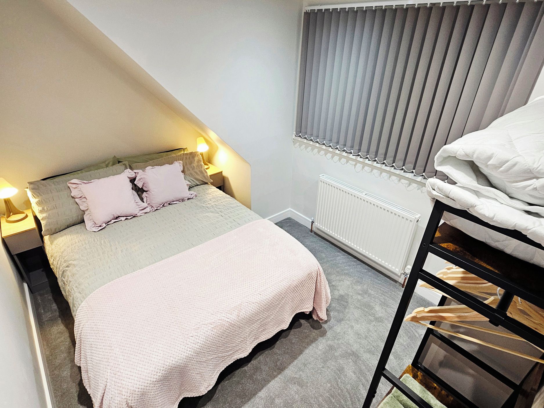 A bedroom with a bed, bedside lamps, a radiator, and a window with blinds.